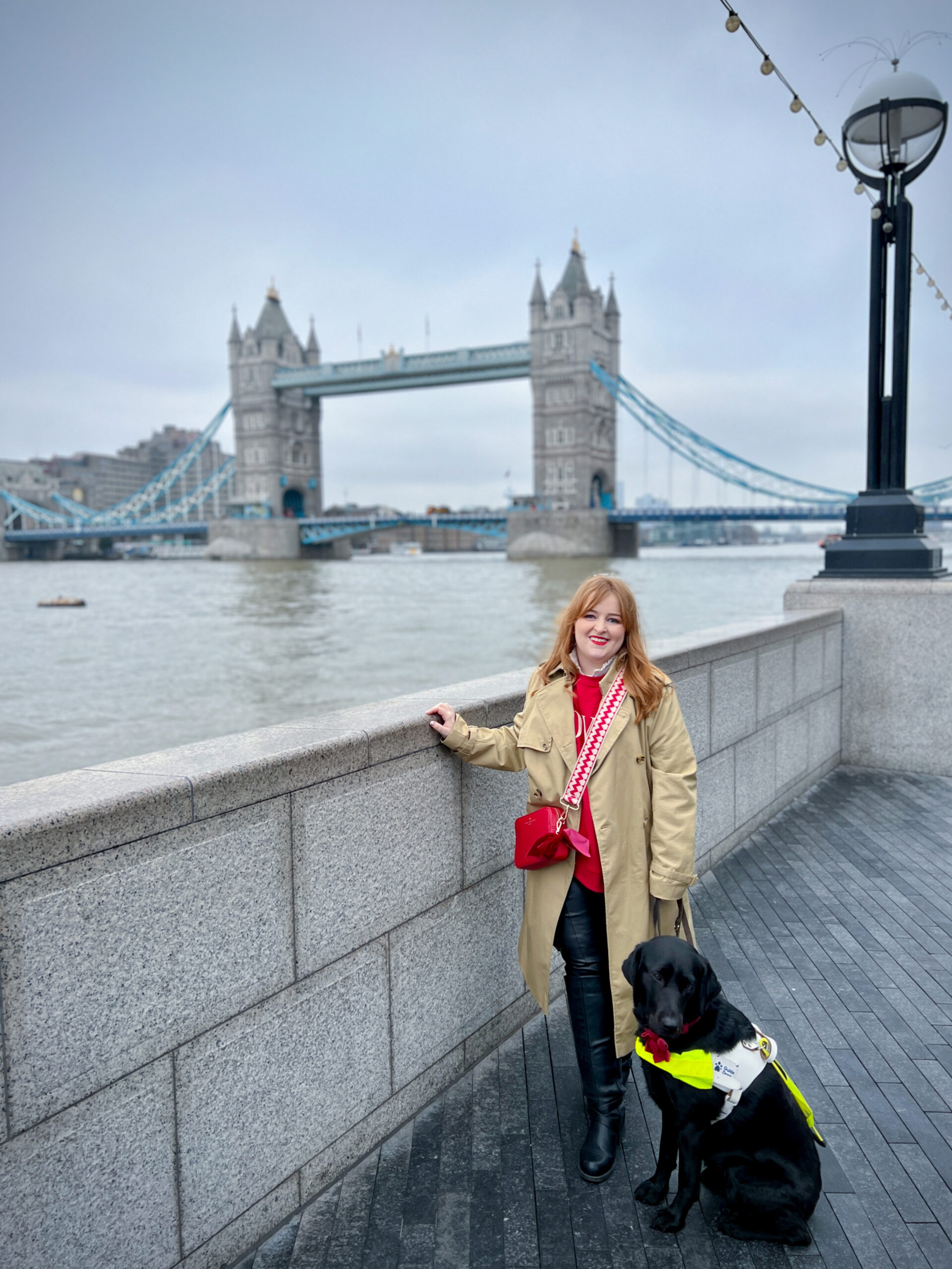 A photo of Emily and guide dog Rosie (a black Labrador) standing in front of Tower Bridge in London