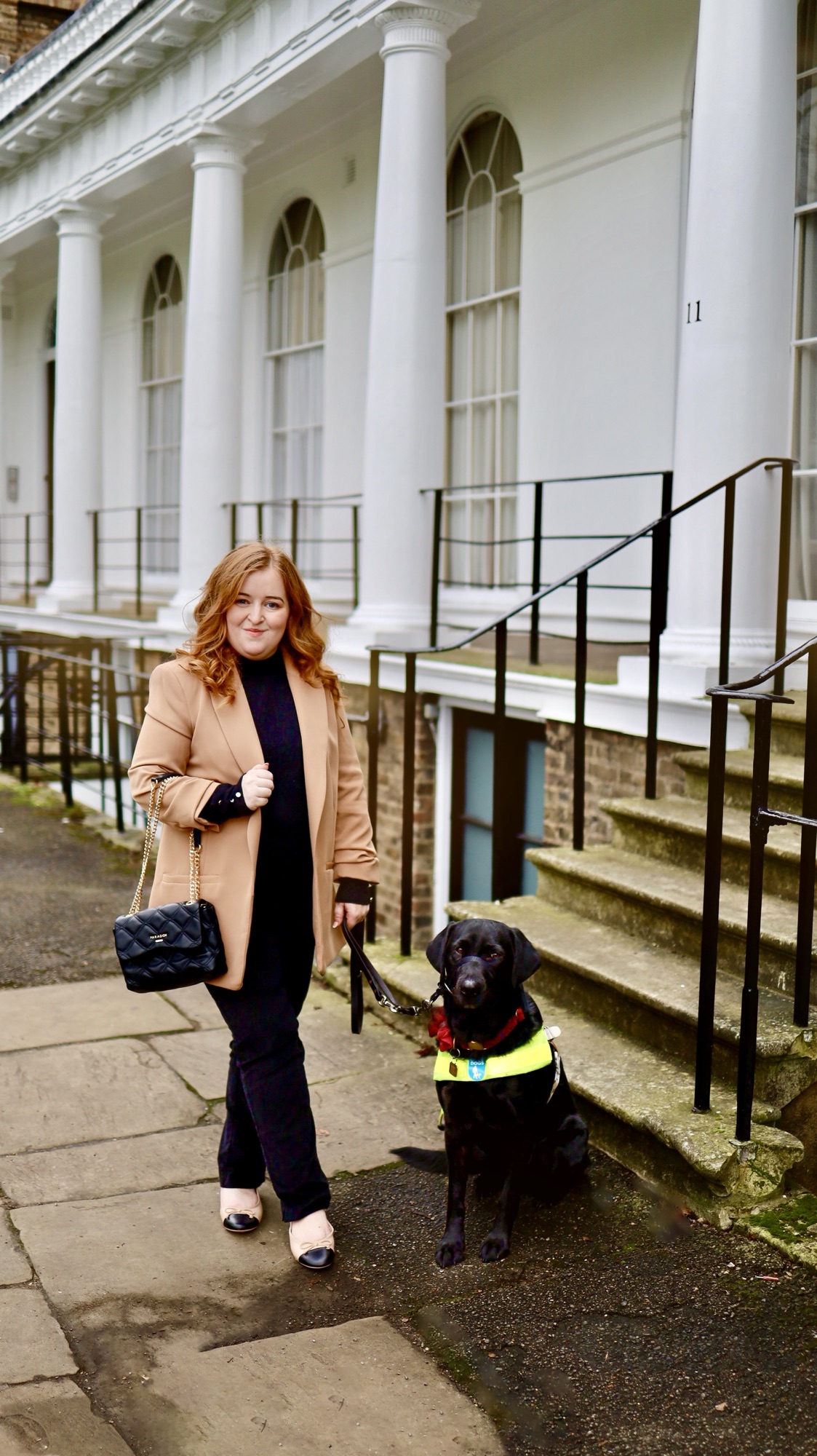 A photo of Emily wearing a cream blazer, black jeans and black roll neck jumper standing with her guide dog Rosie in a Georgian street in London with black railings behind her.