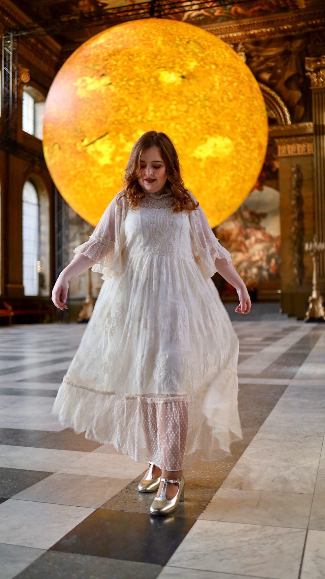 A photo of Emily wearing a white lace maxi dress standing in The Painted Hall in front of Helios.