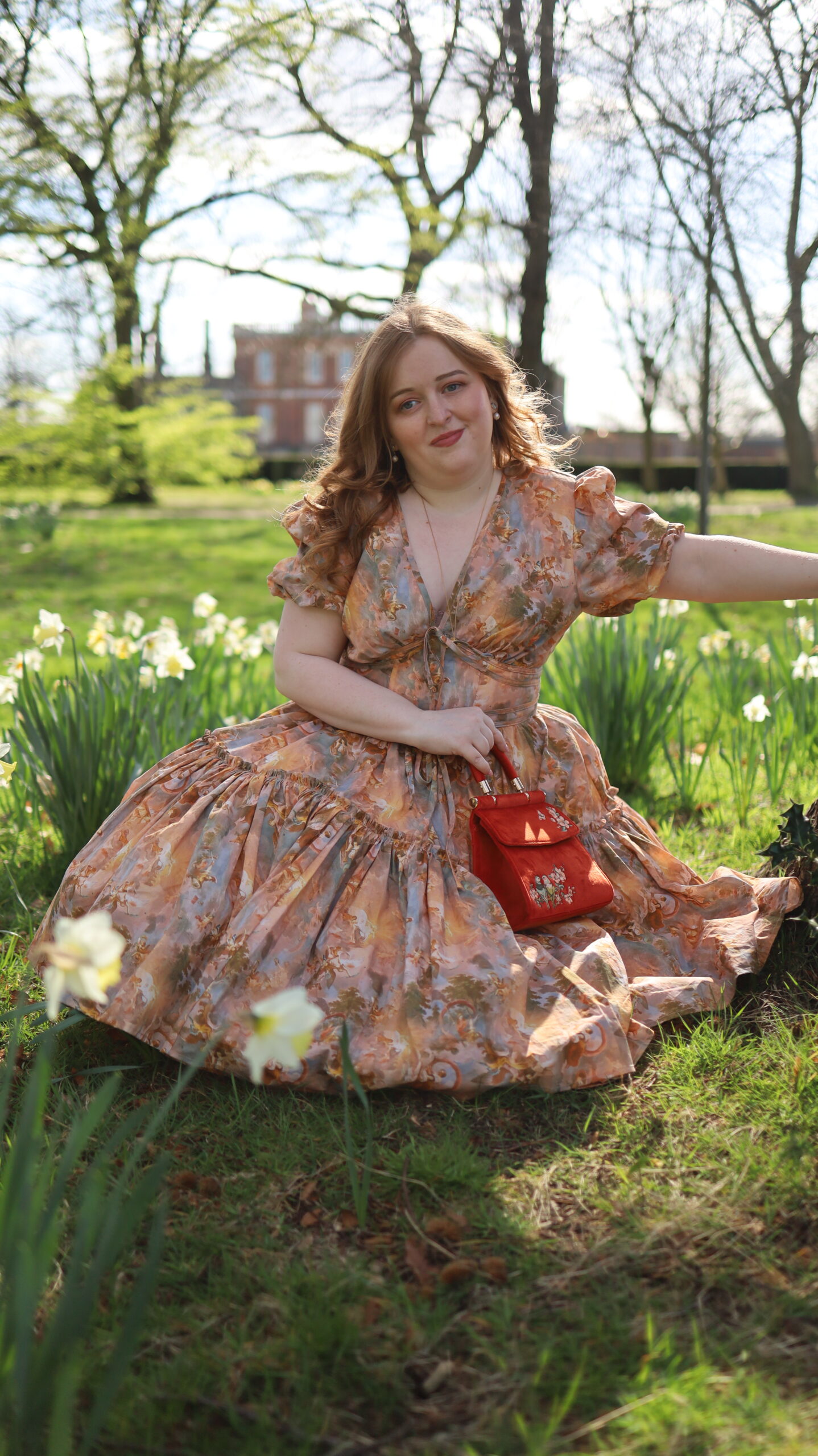 She sits gracefully among blooming daffodils, bathed in the soft light of a spring afternoon. The voluminous skirt of the Ode to Jove dress fans around her like petals, revealing the intricate print in warm tones of gold, peach, and blue. With her head slightly bowed and a gentle smile on her lips, she looks every bit the picture of pastoral elegance—like a character from a timeless fairytale.