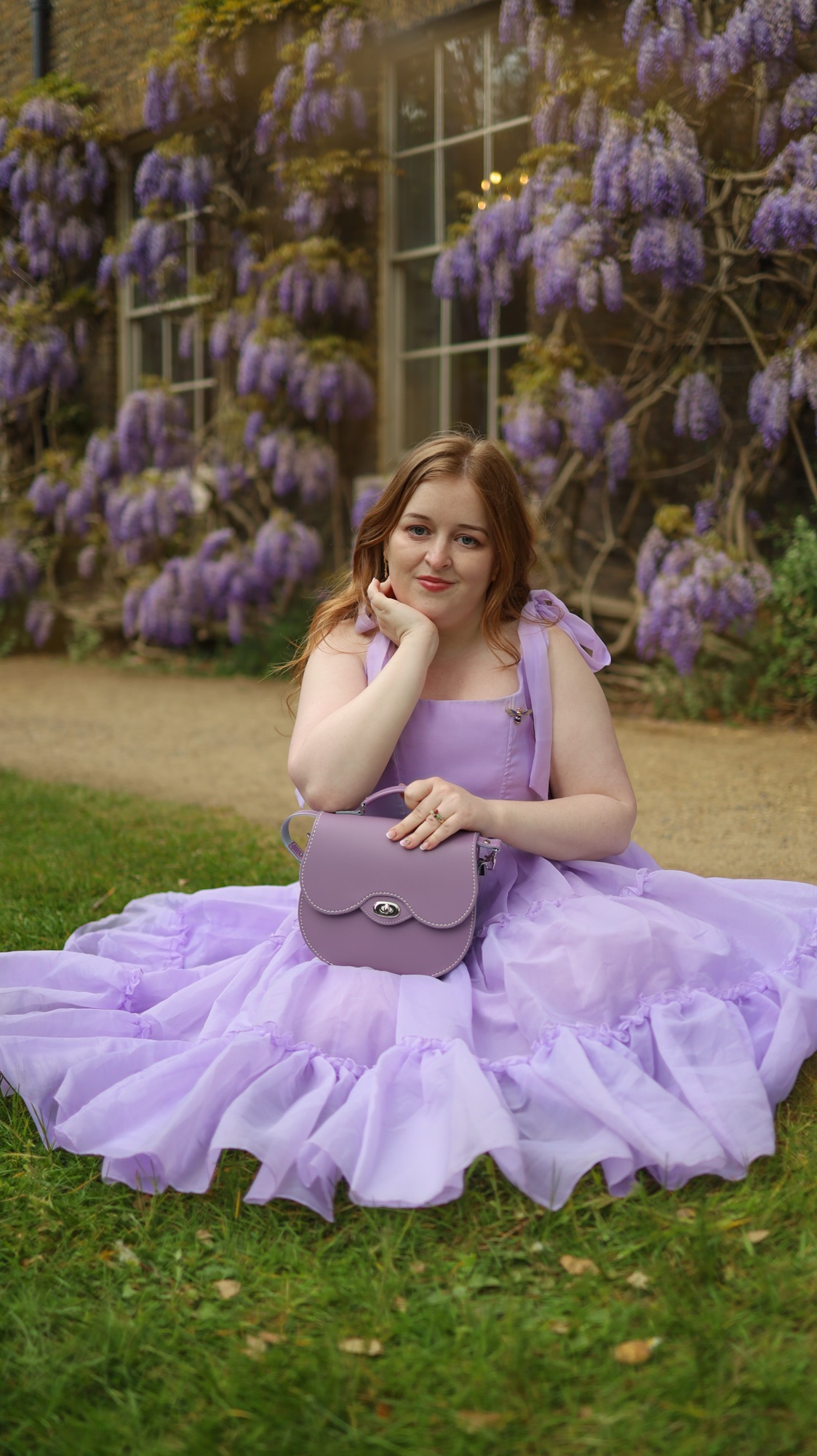 The same woman sits on the grass in front of a wisteria-covered building. Her dress fans out around her like petals, and she rests her face on her hand with a soft smile.