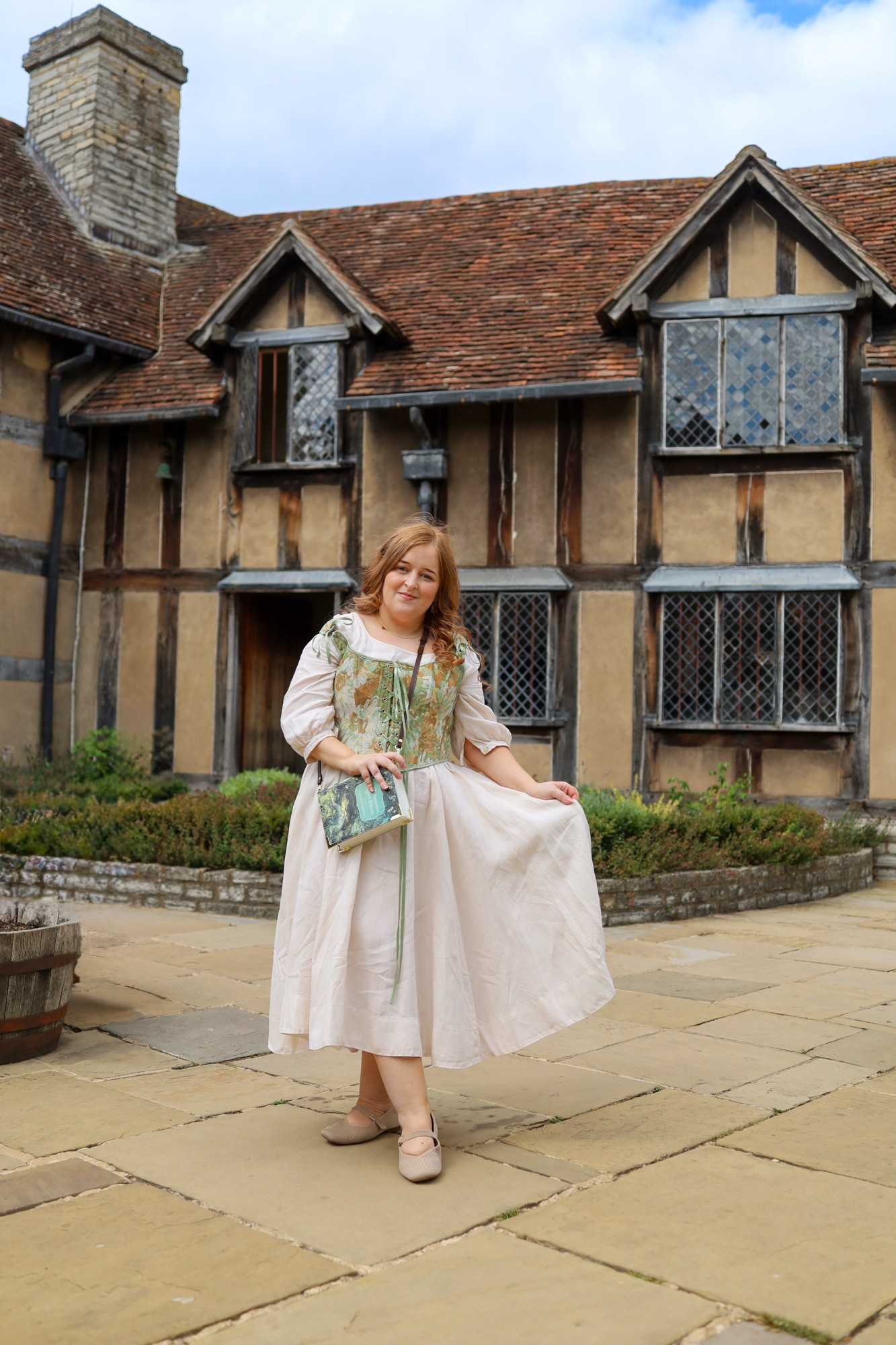 A woman in a light dress poses, holding part of her skirt, standing on a stone path in front of a historic, timber-framed house with diamond-paned windows and a tiled roof.